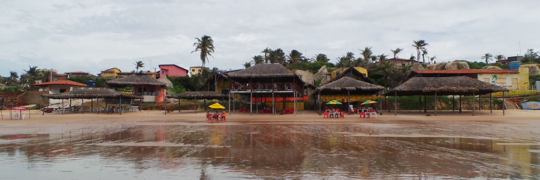 Barracas de palha e mesas na areia da Praia de Quixaba, com casas coloridas e coqueiros ao fundo